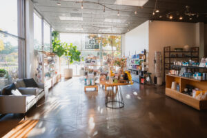 A detailed view of the interior retail and reception area of Changes Salon & Day Spa. The wide shot shows the bright, sunlit space with floor-to-ceiling windows, polished concrete floors, and multiple product display shelves filled with beauty items. A central round table features a yellow arrangement, and a gray seating area is visible on the left, capturing the inviting atmosphere of the East Bay salon.